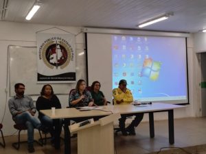 Pessoas sentadas em volta de uma mesa. Uma mulher ao centro de mesa ao público que está a sua frente