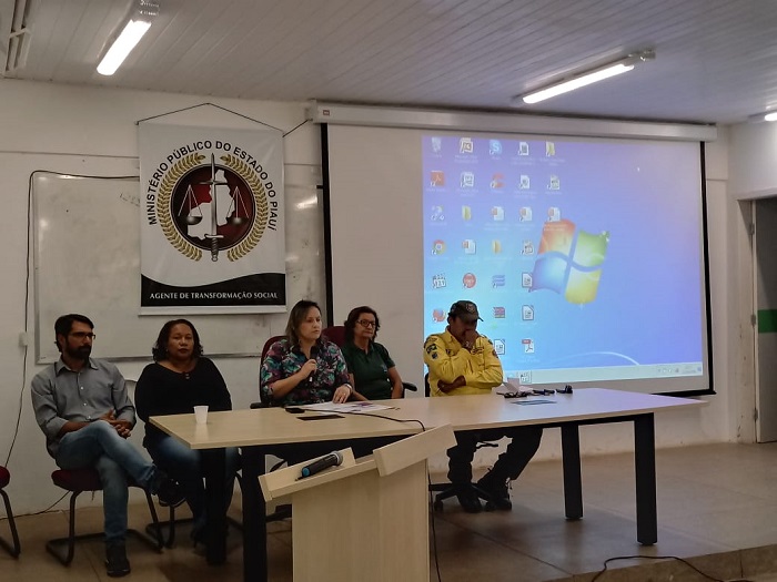 Pessoas sentadas em volta de uma mesa. Uma mulher ao centro de mesa ao público que está a sua frente