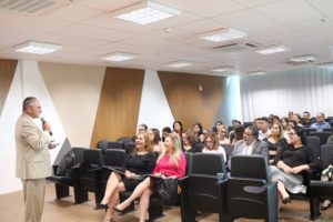 Pessoas sentadas em auditório observando homem que está de pé falando com microfone nas mãos