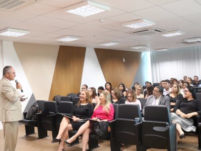 Pessoas sentadas em auditório observando homem que está de pé falando com microfone nas mãos