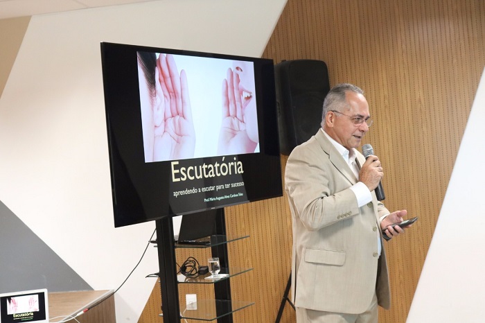 Homem de pé falando para o público. Ao fundo uma tela com a palavra: escutatória