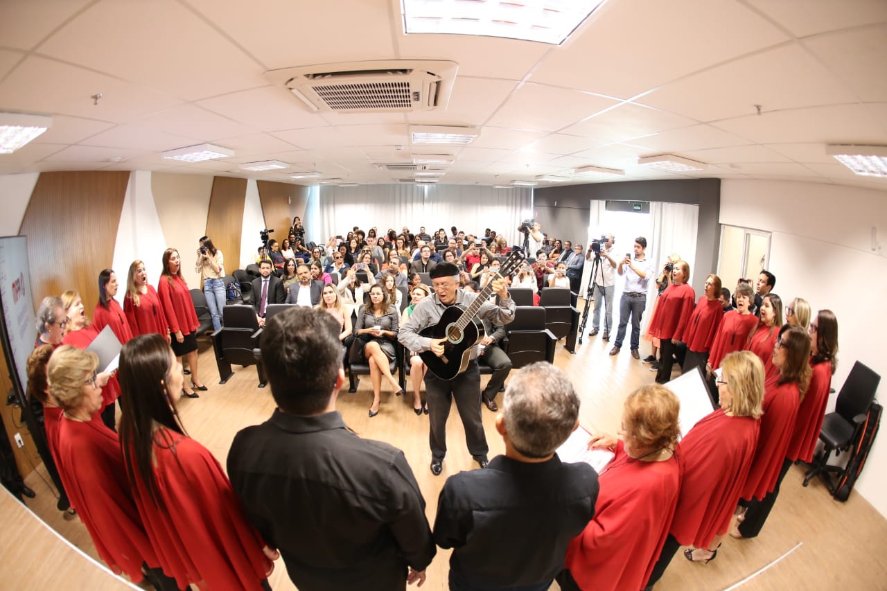 Coralistas em primeiro plano cantam enquanto maestro toca violão. Ao fundo, público aprecia o coral. 