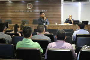 Sala com um homem em pé e várias pessoas sentadas prestando atenção