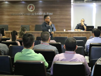 Sala com um homem em pé e várias pessoas sentadas prestando atenção