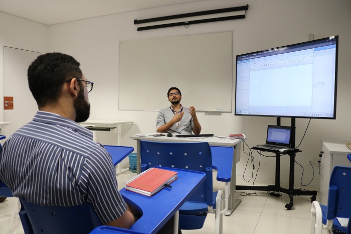 Pessoa sentada em sala de aula. A pessoa observa o instrutor que a sua frente fala para os demais pessoas que estão na sala. 