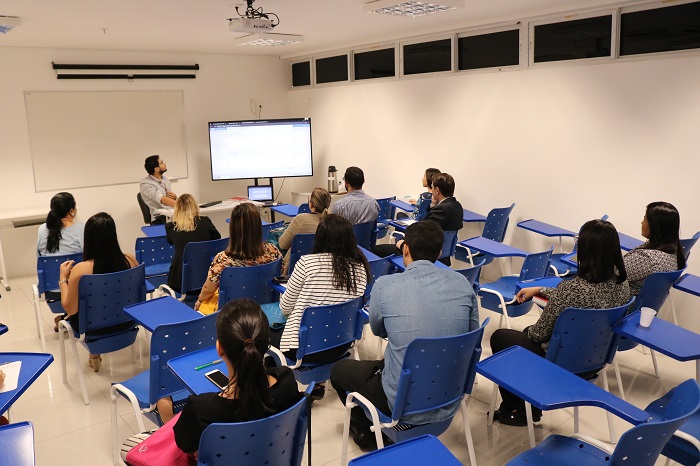 Pessoas sentadas em sala de aula assistindo treinamento. Ao fundo há um homem sentado em uma mesa ao lado de uma grande tela por meio da qual mostra as informações do treinamento 