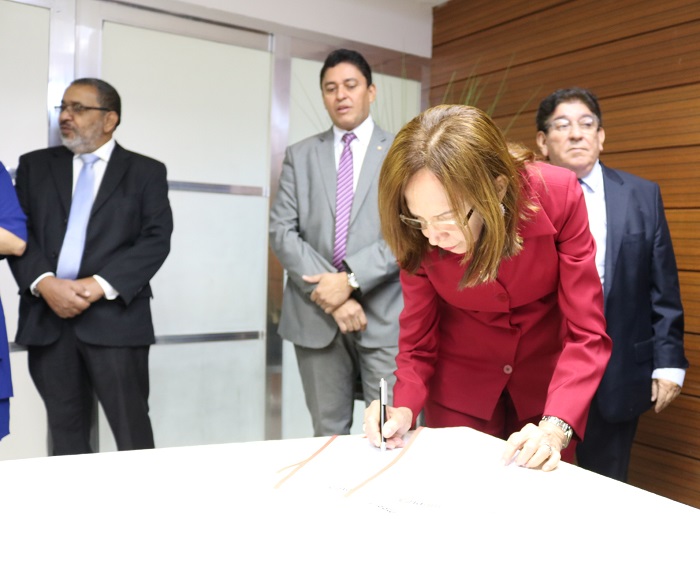 Pessoa assinando documento sobre uma mesa. Ao fundo pessoas de pé a observam 
