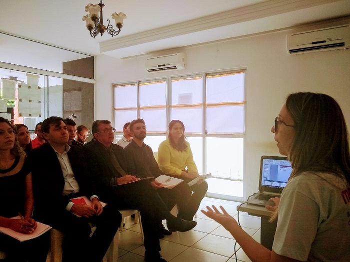 Mulher de pé gesticulando e um grupo de pessoas ao fundo sentadas