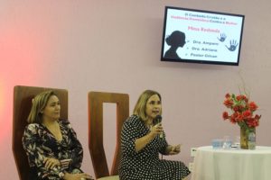 Duas pessoas sentadas. Uma das pessoas segura um microfone enquanto fala com o público