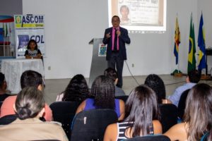Pessoas sentadas em auditório. Ao fundo há um homem que está de pé falando para a plateia