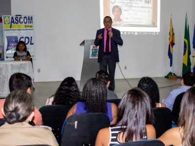 Pessoas sentadas em auditório. Ao fundo há um homem que está de pé falando para a plateia