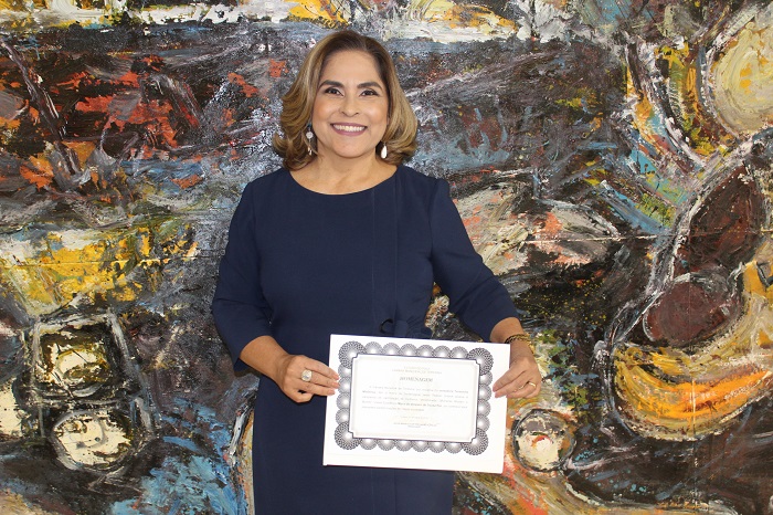 Mulher de pé sorrindo e segurando com as duas mãos um diploma da homenagem recebida 