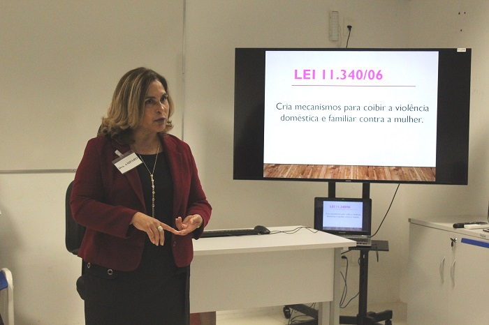 Mulher de pé falando paras as pessoas que a observam. Ao fundo há um televisão com slides sendo exibidos 