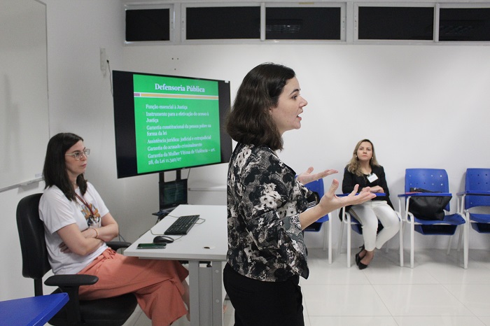 Três mulheres. A primeira está de pé gesticulando com as mãos. A segunda está sentada logo atrás da primeira e a terceira mulher está sentada ao fundo da sala 