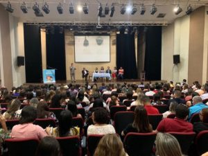 Pessoas sentadas em auditório. A fundo, no palco pessoas sentadas atrás de uma mesa observando um home que de pé fala para plateia