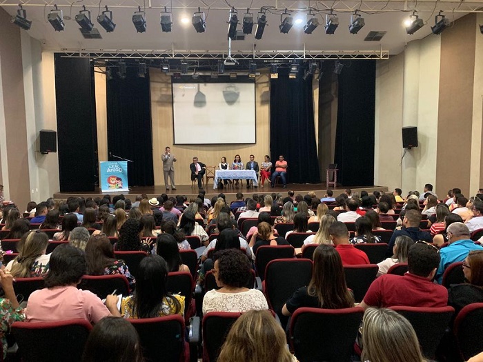 Pessoas sentadas em auditório. A fundo, no palco pessoas sentadas atrás de uma mesa observando um home que de pé fala para plateia