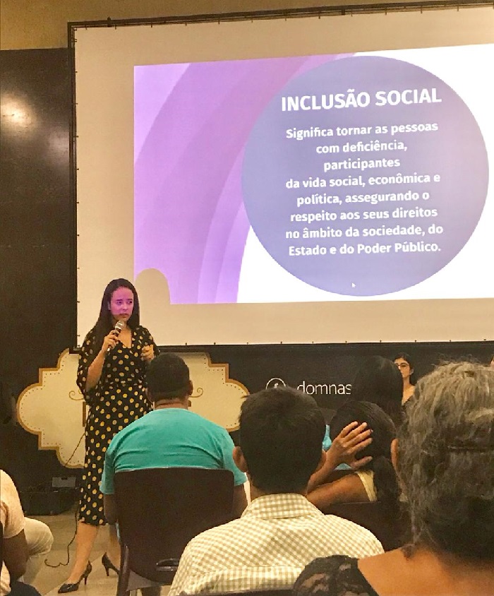 Mulher de pé falando com pessoas que estão sentadas a sua frente 