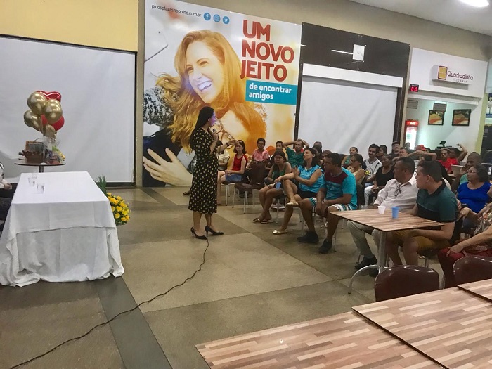 Pessoas sentadas em auditório observando uma mulher que está de pé falando 