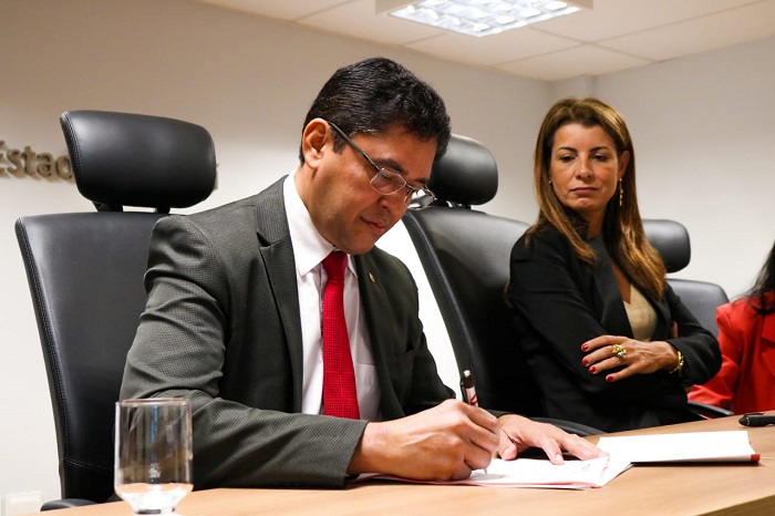 Pessoa assinando documento sobre uma mesa. Ao fundo pessoas de pé a observam 