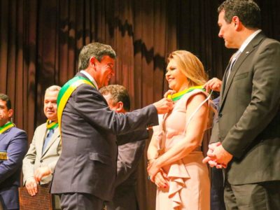 Momento que o Governador do Piauí, Wellington Dias, entrega medalha para a PGJ Carmelina moura. Ela sorri e olha para frente enquanto recebe a honraria.