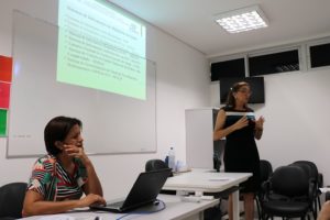 Uma mulher em pé falando e uma mulher sentada ao computador