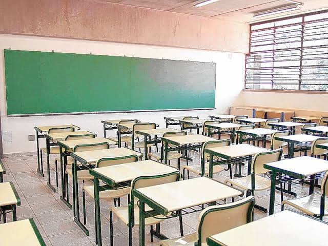 Quadro de giz ao fundo e carteiras enfileiradas em sala de aula