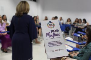 Em primeiro plano, panfleto sobre o PRO MULHER. Ao fundo, pessoas sentadas