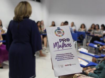 Em primeiro plano, panfleto sobre o PRO MULHER. Ao fundo, pessoas sentadas