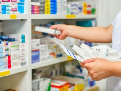 Fotografia que mostra as mãos de um profissional de saúde, arrumando caixas de medicamentos em uma estante