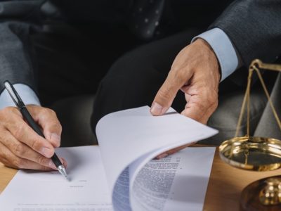 Imagem da mão de uma pessoa assinando um documento sobre uma mesa. Ao lado há uma balança representando a Justiça