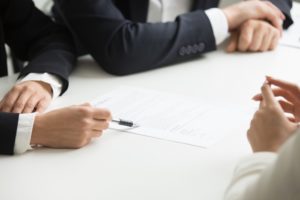 Imagem de mãos de pessoas sobre uma mesa. Um das pessoas está com uma caneta e aponta para um documento que está sobre a mesa